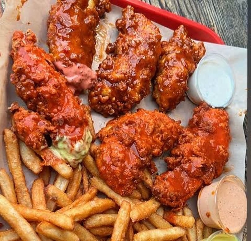 Close-up of a tray of crispy, saucy chicken tenders with fries and dipping sauces.