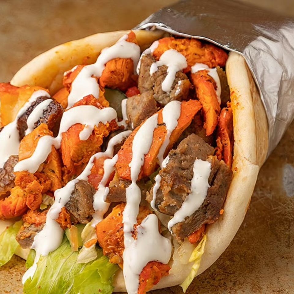 A pita bread wrap filled with chicken, beef, lettuce, and white sauce, partially wrapped in foil.