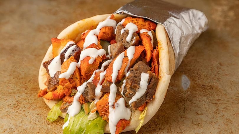 A pita bread wrap filled with chicken, beef, lettuce, and white sauce, partially wrapped in foil.