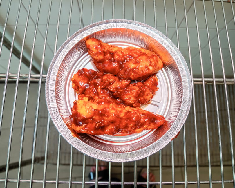 Three saucy chicken tenders in a foil pan on a metal rack, viewed from above.