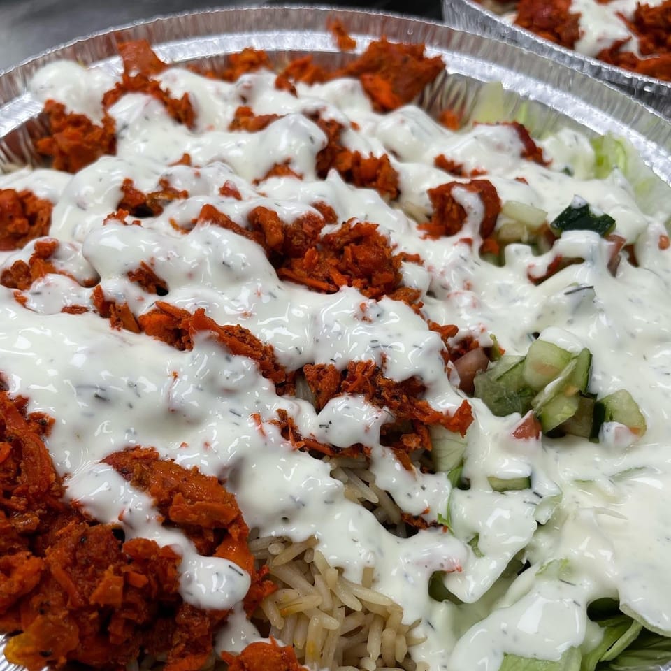 Close-up of a foil container filled with rice, red chicken, salad, and white sauce.