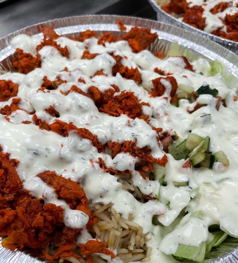 Close-up of a foil container filled with rice, red chicken, salad, and white sauce.