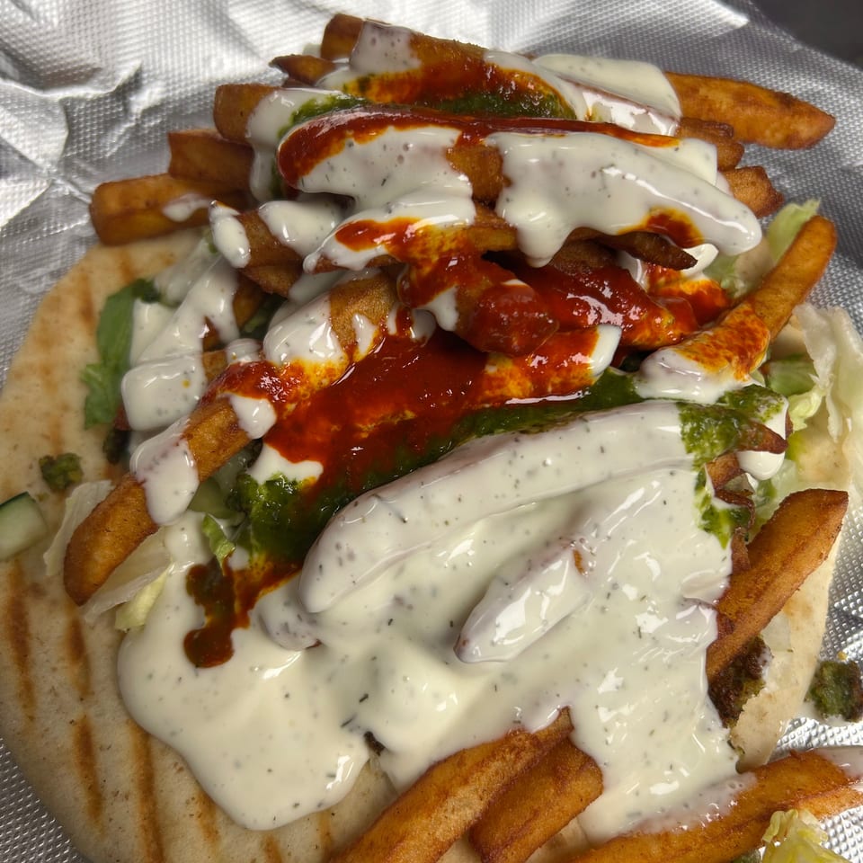 A pita bread topped with french fries, lettuce, white sauce, and red and green sauces.