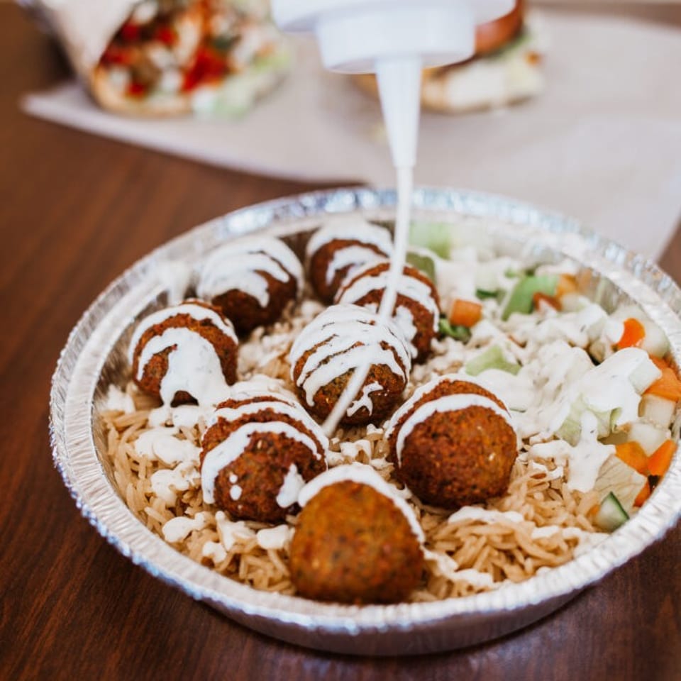 A squeeze bottle drizzles white sauce over falafel, rice, and salad in a foil container.