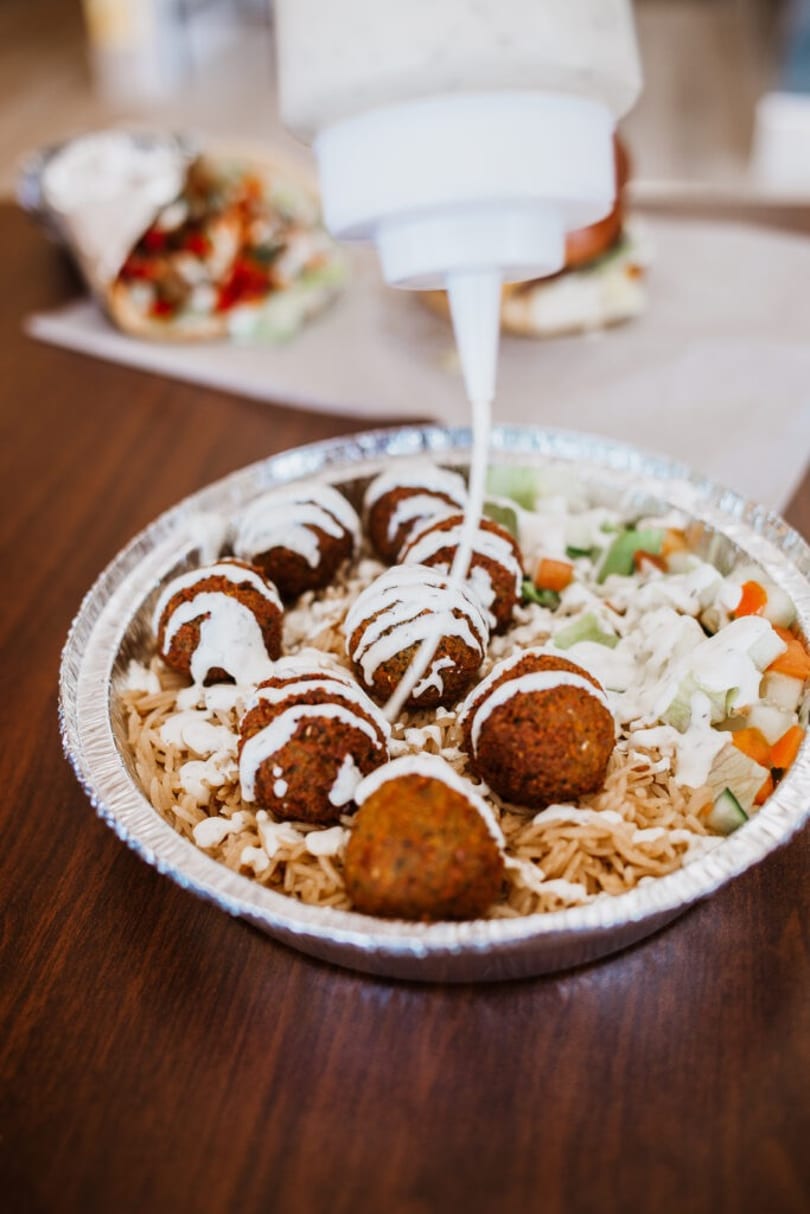 A squeeze bottle drizzles white sauce over falafel, rice, and salad in a foil container.
