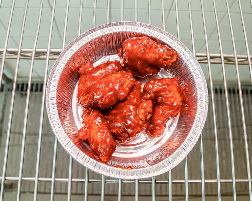 Spicy red chicken wings in a foil container, resting on a metal rack.