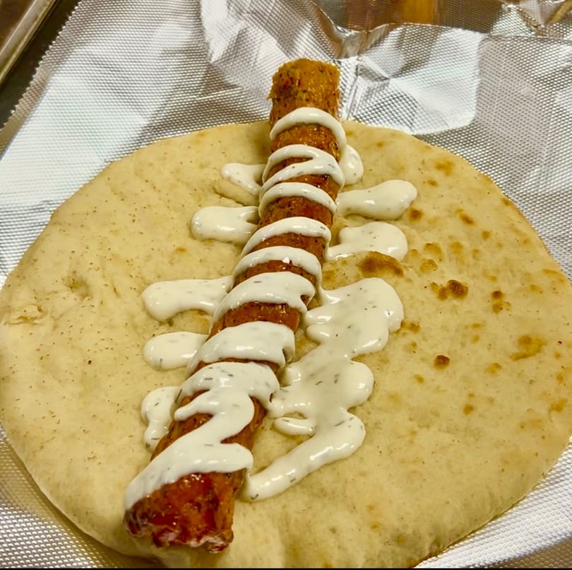 Grilled kebab skewer with white sauce on a flatbread, served on aluminum foil.